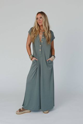 A person with long blonde hair is standing against a plain background, donning a loose-fitting Three Bird Nest Logan Wide Leg Jumpsuit in a light olive shade. The short-sleeved jumpsuit features buttons down the front. They have their hands in the pockets and wear multiple bracelets along with beige slip-on sandals.