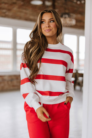 A person with long hair wears the Callahan Striped Pullover and Joggers Set by Liam & Company, standing in a room with large windows. With one hand in their pocket, they glance to the side, slightly smiling.