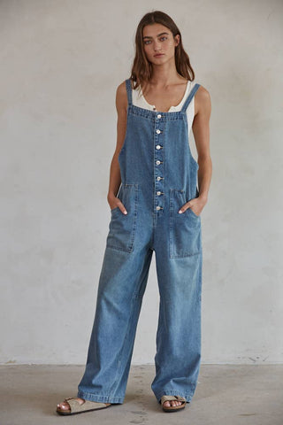 A person stands against a plain backdrop, wearing By Together's Kodi Denim Square Neck Button-down Wide Leg Overalls over a white tank top. They have their hands in the overall pockets and are wearing open-toed sandals. They have shoulder-length hair and a neutral expression.