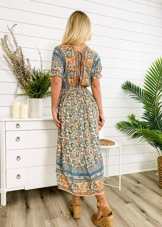 A woman with long blonde hair wearing the Finn Tie Back High Low Dress by Anna Kaci Wholesale stands with her back to the camera in a white room adorned with plants and a dresser. She is sporting wedge sandals, and the summer dress showcases intricate blue, orange, and cream designs.