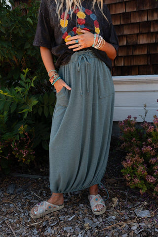 A person wearing a dark t-shirt with a colorful floral design and the Rylie Drawstring Maxi Skirt - Teal Boho Skirt by Three Bird Nest stands outdoors. They accessorize with numerous colorful bracelets, rings, and a necklace, embodying the boho western wardrobe. The person is also wearing woven sandals.