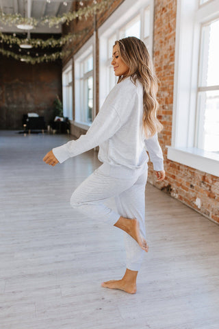 A woman with long, wavy hair stands on one leg in a cozy, sunlit room with exposed brick walls and large windows. She wears an Alison Half Zip Loungewear Set by Liam & Company, made from luxurious fabric. Barefoot and appearing relaxed and serene, she basks in the bright, open space.