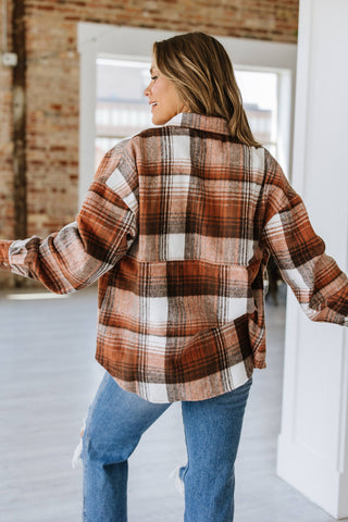 Standing indoors with long hair and facing away, the person is wearing the fashionable Kelsea Plaid Shacket in brown, white, and orange from Liam & Company. They have paired it with blue jeans against a backdrop of a brick wall and large windows.