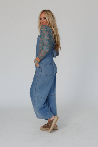 Wearing the Three Bird Nest Dock Town Button Up Jumpsuit in denim with a blue lace top underneath, a person with long blonde hair stands sideways with one hand in their pocket. Sporting brown sandals and several colorful bracelets on their wrist, they smile while looking over their shoulder.