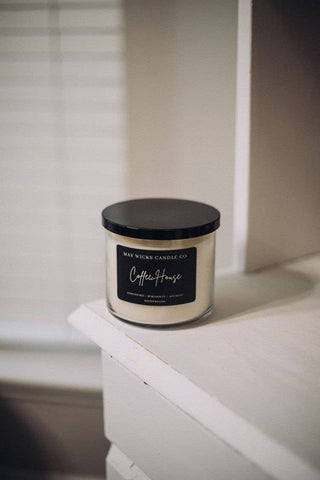 A white candle in a clear glass jar with a black lid sits on a white wooden shelf. The label on the jar reads "Mav Wicks Candle Co. Mahogany Teakwood Candle: 17 OZ," known for its soy-based wax and sophisticated fragrance. In the background, blurred vertical blinds are visible.