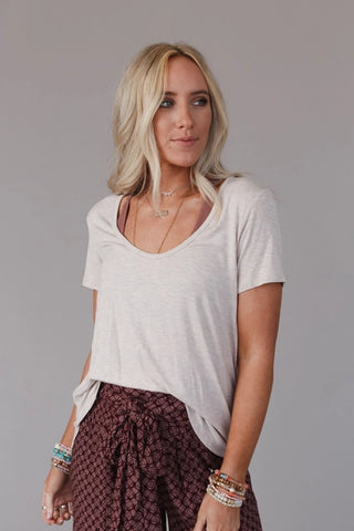 A woman with long blonde hair is wearing a Three Bird Nest Moxen Scoop Neck Bralette Tee and patterned high-waisted pants. She has several bracelets on her right wrist and is standing against a plain, light gray background.