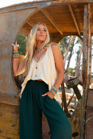 A person with long, blonde hair stands against a weathered, rusted structure, gazing off into the distance. They are wearing the Elara Top in Neutral by Three Bird Nest and dark wide-leg pants, accessorized with bracelets and necklaces that reflect a boho style. Tall trees are visible in the background.