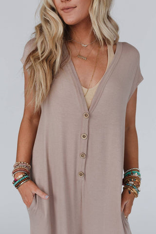 A woman with long, wavy blonde hair wears the loose-fitting Kendrick Mocha Jumpsuit by Three Bird Nest, featuring buttons down the front. She accessorizes with layered necklaces and colorful bracelets, her hands casually placed in the jumpsuit pockets. The background is plain and light-colored.