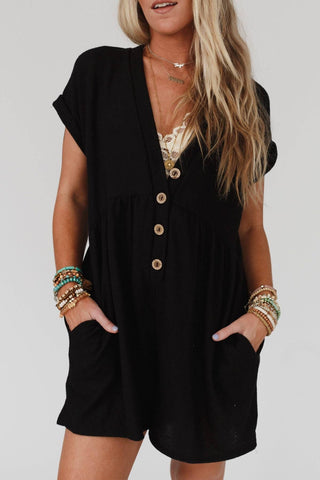 A woman with long blonde hair wearing the Sage and Wildflowers Romper in black by Three Bird Nest, a comfortable button-up romper with short sleeves and pockets. She accessorizes with layered necklaces and multiple bracelets on both wrists, adding a boho-chic touch. The background is plain and light-colored.