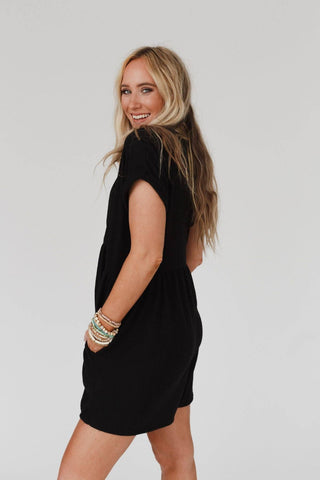 A woman with long blonde hair is smiling and wearing the Sage and Wildflowers Romper - Black by Three Bird Nest. She is turned slightly to the side, showcasing the back and side of her outfit. Her left arm is adorned with multiple colorful bracelets. The background is plain and light-colored.