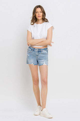 A woman stands against a plain, white background with her arms crossed. She is wearing a white T-shirt, white sneakers, and the Kylie Mid Rise Raw Distressed Hem shorts from VERVET by FLYING MONKEY made from comfortable stretch denim. She has long, wavy hair and a neutral expression.