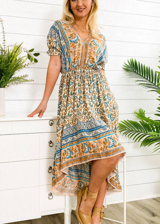 A person wearing the vibrant Finn Tie Back High Low Dress by Anna Kaci Wholesale stands indoors against a white paneled wall. They're leaning against a white chest of drawers, surrounded by potted plants. They accessorize with light-colored sandals featuring ankle straps.