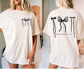 In a softly lit room with a bed, someone confidently wears Western Edge Boutique's oversized tee, 'She bows down for no one,' featuring black line art bows on the back—an embodiment of self-love and quiet royalty.