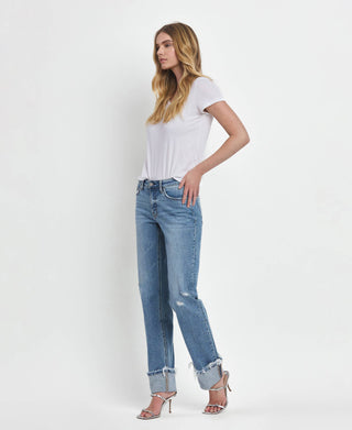 A woman with long blonde hair is standing against a plain white background. She is wearing a white T-shirt, Lovervet by VERVET's Dezie Mid Rise Cuffed Straight Jeans All Sizes 1-24 in soft stretch denim, and silver open-toed high heels. Her right hand rests on her hip as she gazes off to the side.