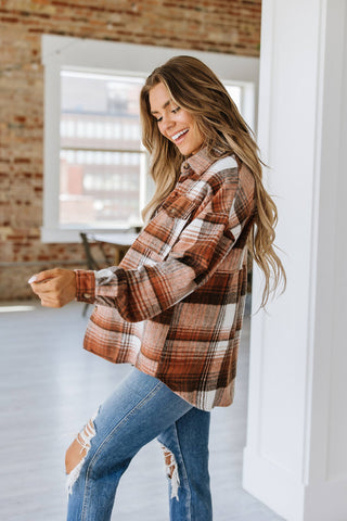 A smiling woman with long hair stands in a well-lit room featuring exposed brick walls. She is wearing the trendy Kelsea Plaid Shacket by Liam & Company and a pair of ripped jeans, looking downward with a relaxed expression.