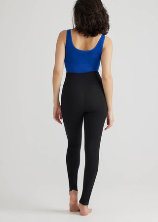 A person with shoulder-length dark hair is standing barefoot on a plain background, facing away from the camera. They are wearing a blue sleeveless top and Yummie's Grady Shaping Legging with Side Pockets in Black, made from cotton stretch for added convenience.