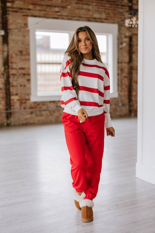 A long-haired person smiles in a Liam & Company Callahan Striped Pullover and Joggers Set while wearing furry boots, standing on a light wood floor in a bright room with brick walls and large windows.