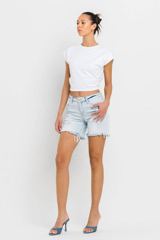 A person with their hair tied back stands against a white background, wearing a white cropped T-shirt, light blue Kolson Mid Rise Distressed Stretch Denim Shorts by VERVET by FLYING MONKEY, and blue open-toe heels. Their left hand is on their hip, while their right arm rests by their side.