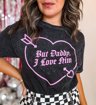 A person wears Mugsby's "But Daddy I Love Him" Valentine's Shirt featuring a pink heart and Gothic script. It's paired with a black-and-white checkered skirt amidst silver and pink decorations, creating a classic country song vibe.