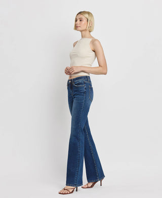 A woman with short blonde hair is standing against a plain white background. She is wearing a sleeveless white top, Kip Mid Rise Relaxed Bootcut Jeans from Lovervet by VERVET, and black high-heeled sandals. She is facing slightly to the left with her hands resting by her sides.