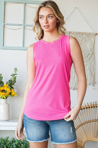 A woman with wavy blonde hair is wearing a bright pink Georgia Curvy Ribbed Tank Top from Western Edge Boutique and denim shorts. She stands indoors in front of a wall with window pane decor and a macramé hanging. A vase with yellow flowers is on a table next to a green plant.