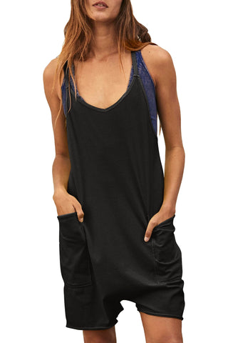 A person with long, wavy hair wears a comfy, loose-fitting Charmo Kaylin Hot Shot Short Romper With Pockets. The black romper is sleeveless and features deep front pockets, revealing a blue tank top underneath. With their hands in the pockets, they stand against a plain white background.