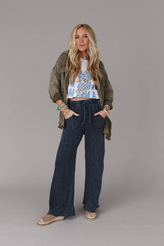 A woman with long blonde hair stands confidently against a neutral background. She wears a green jacket over a white graphic t-shirt, Three Bird Nest's Cooper Relaxing Robin Wide Leg Pant in navy blue with a vintage fade, and beige shoes. Her hands are in her pockets, showcasing bracelets on her wrists.