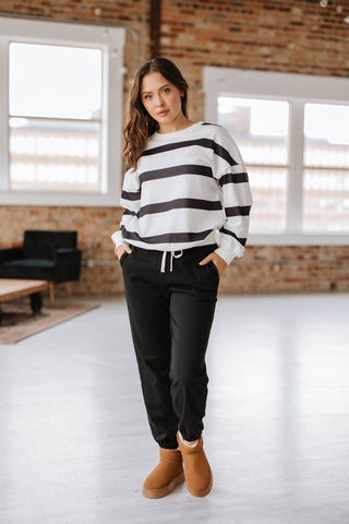 A woman stands indoors on a light floor, wearing the Callahan Striped Pullover and Joggers Set by Liam & Company. Her hands are in her pockets, and she's styled with brown boots. Brick walls and large windows enhance the cozy atmosphere.