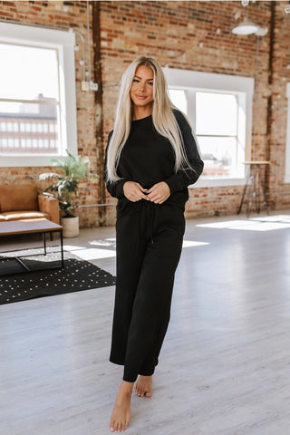 A woman with long blonde hair is standing barefoot in a spacious room with exposed brick walls and large windows, wearing an Alex Black Lounge Set by LIAM AND CO, showcasing both comfort and style. There is a brown couch, a potted plant, and a rug in the background. The room is well-lit with natural light.