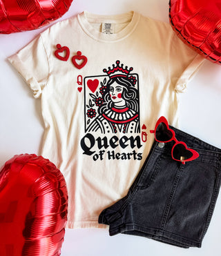 A cream-colored "Queen of Hearts" graphic tee from Mugsby is styled with black denim shorts, heart-shaped sunglasses, and earrings. Surrounded by red heart-shaped balloons, the ensemble is perfect for a whimsical Valentine's Day vibe.