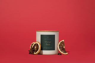 A white soy wax candle from Hico Candle Co., labeled "Orange Spice" in dark green, with a wooden lid, sits on a red background. Two dried mandarin slices and star anise beside it hint at its warm orange spice scent.