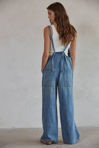 A person with long brown hair is standing and facing away from the camera, wearing a white sleeveless button-down top and By Together's Kodi Denim Square Neck Button-down Wide Leg Overalls. The overalls feature large back pockets and strap details, set against a plain, light-colored wall.