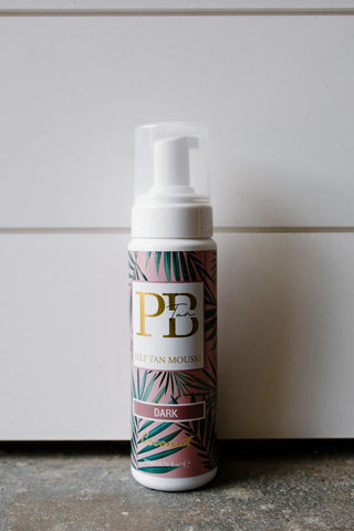 A bottle of Western Edge Boutique's Self Tanner Mousse - Dark is set against a light gray background. The vegan-friendly formula, enriched with EcoCert DHA, promises a natural glow. The bottle has a pump dispenser and is decorated with green leaves on a pink background, labeled "Dark.