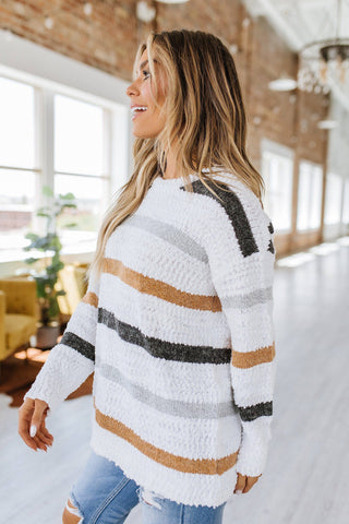 A woman with long blonde hair is standing indoors, wearing a cozy Mckinley Striped Popcorn Sweater by Liam & Company and ripped light blue jeans. She is looking to the side and smiling, with a bright, airy room and large windows in the background. This stylish addition is perfect for any cool-weather wardrobe.