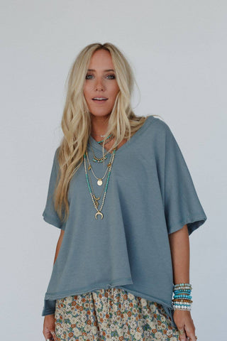 A person with long blonde hair is standing against a plain background, wearing the Payton Top from Three Bird Nest and a bohemian-style floral skirt. Their layered necklaces feature various pendants, and they have several bracelets on their left wrist, looking directly at the camera.