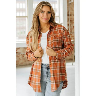 A person with long hair stands indoors, exuding style from Western Edge Boutique. They're wearing a white top beneath the Palmer Oversized Flannel and blue jeans. Smiling as they adjust their shirt, the relaxed fit complements the windows and brick wall behind them.