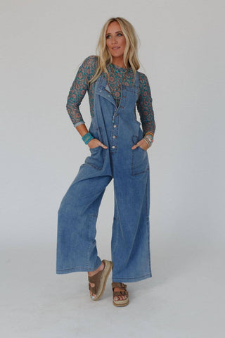 A woman with long blonde hair is posing against a plain background. She is wearing a patterned long-sleeve top under the Three Bird Nest's Dock Town Button Up Jumpsuit - Denim and open-toe platform sandals. With one hand in her pocket, she looks to the side showcasing her boho style clothing effortlessly.