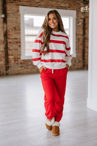 A person with long hair smiles brightly in a room with large windows and a brick wall, wearing the comfortable Callahan Striped Pullover and Joggers Set by Liam & Company, paired with red pants and brown boots lined with white fur for an effortlessly stylish look.