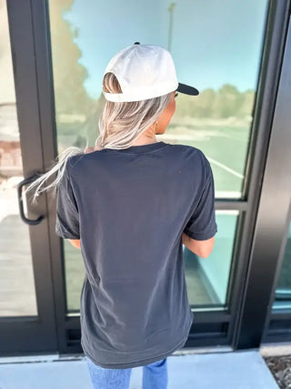 A person with long blonde hair, wearing a Bird & Co Wholesale Tees' Little Ray Of Black Graphic Tee in large, jeans, and a white cap, stands facing a glass door. The scene is outdoors with a sunny reflection in the background.