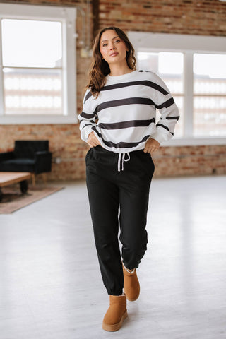 In a well-lit room with large windows and exposed brick walls, a person wears the comfortable Callahan Striped Pullover and Joggers Set from Liam & Company, paired with tan boots.