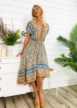 A woman stands in a room adorned with plants, wooden flooring, and white walls. She is wearing the Finn Tie Back High Low Dress by Anna Kaci Wholesale, featuring blue and orange floral prints. She leans on a white dresser beside a white candle and a potted arrangement. She completes her look with tan shoes.