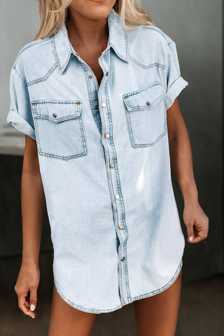 A person is wearing the Aapparella Vintage Light Wash Flap Pockets Rounded Hem Shirt with short sleeves casually rolled up. The shirt features button closures down the front and two breast pockets with flaps. The minimalistic and slightly blurred background highlights the outfit.