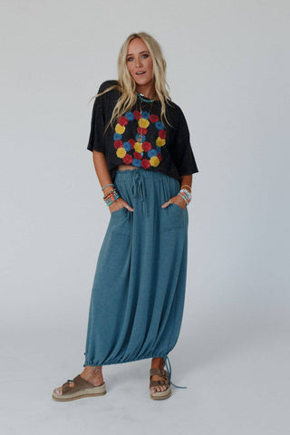 A woman with long blonde hair is standing against a plain background. She is wearing a black t-shirt with colorful circular designs, the "Rylie Drawstring Maxi Skirt - Teal Boho Skirt" by Three Bird Nest featuring an elastic drawstring waistline, bracelets, necklaces, and brown sandals. With her hands in her skirt pockets, she perfectly embodies a boho western wardrobe vibe.