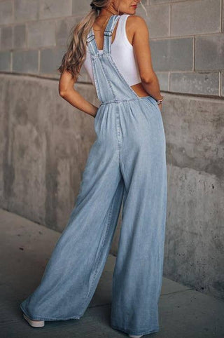 A person with long hair is standing against a concrete wall, facing away from the camera. They are wearing a white sleeveless top and light blue Honeypeach Everson Lightweight Denim Wide Leg Denim Overalls. The person has one hand in a pocket and the other arm relaxed by their side, effortlessly showcasing their fashion overalls.