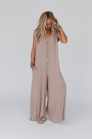 A woman with long blonde hair is standing against a plain background, wearing the Kendrick Mocha Jumpsuit by Three Bird Nest. This loose, sleeveless, beige jumpsuit features buttons down the front. She accessorizes with multiple colorful bracelets on her right wrist and poses with one foot slightly forward, perfectly embodying the boho chic jumpsuit style.