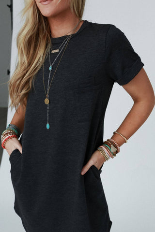 A person with long blonde hair is wearing the "On The Daily Pocket Tee Dress" in charcoal by Three Bird Nest, with hands in the pockets. They are accessorized with several long necklaces and multiple bracelets on both wrists. The background is a simple, neutral indoor setting.