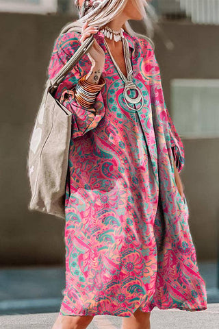 A person wearing the Retro Paisley Tab Sleeve Dress: Rose by Pretty Bash, featuring a bold pink, green, and blue paisley pattern in a colorful, loose-fitting bohemian chic style, stands with a large beige shoulder bag. They have long blonde hair and are accessorized with several bracelets and a large circular pendant necklace.