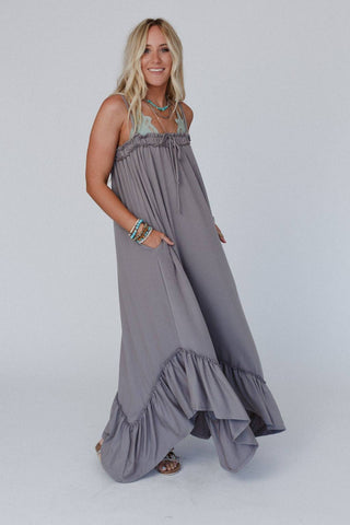 A person stands against a plain background wearing the Heavenly Glow Maxi Dress - Mushroom by Three Bird Nest, which exudes romantic charm with its ruffled accents. They have long, wavy blonde hair and are smiling. They accessorize with several bracelets and wear sandals, with their right hand in their dress pocket.