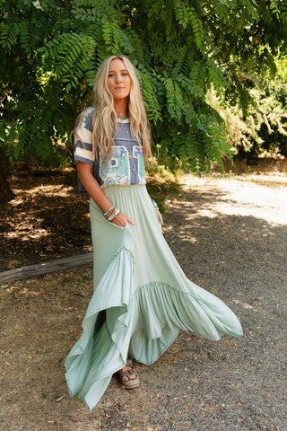 A woman with long blonde hair stands outdoors in partial shade. She is wearing a colorful graphic T-shirt and the Logan Forever Beauty Maxi Skirt in Pale Sage from Three Bird Nest, along with bangles on her wrists. The background features green foliage and patches of sunlight on the ground.