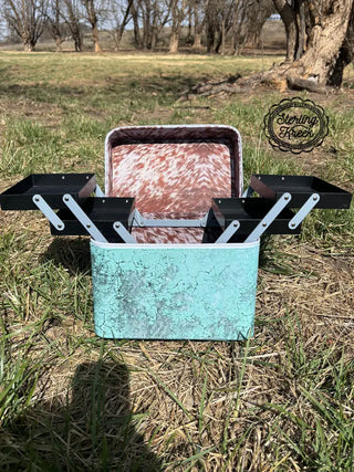 A vintage-style "Heard your Makeup Tool Box" by Western Edge Boutique, featuring a turquoise exterior and marbled interior, sits on the grass in a natural setting. Its rustic design boasts several outward-extending compartments, ideal for organizing makeup tools. In the background, trees and the Western Edge Boutique logo add to the allure of this must-have item for enthusiasts.